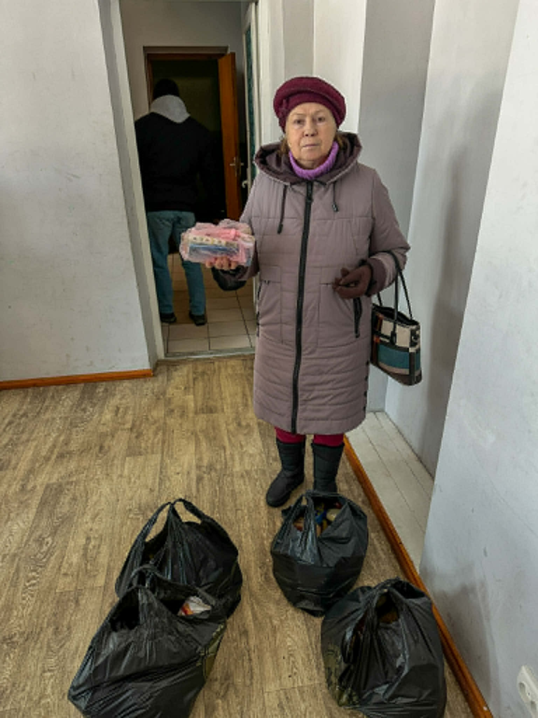 Elderly woman in a mauve winter coat holding a food item, with several filled aid bags placed on the floor in front of her.