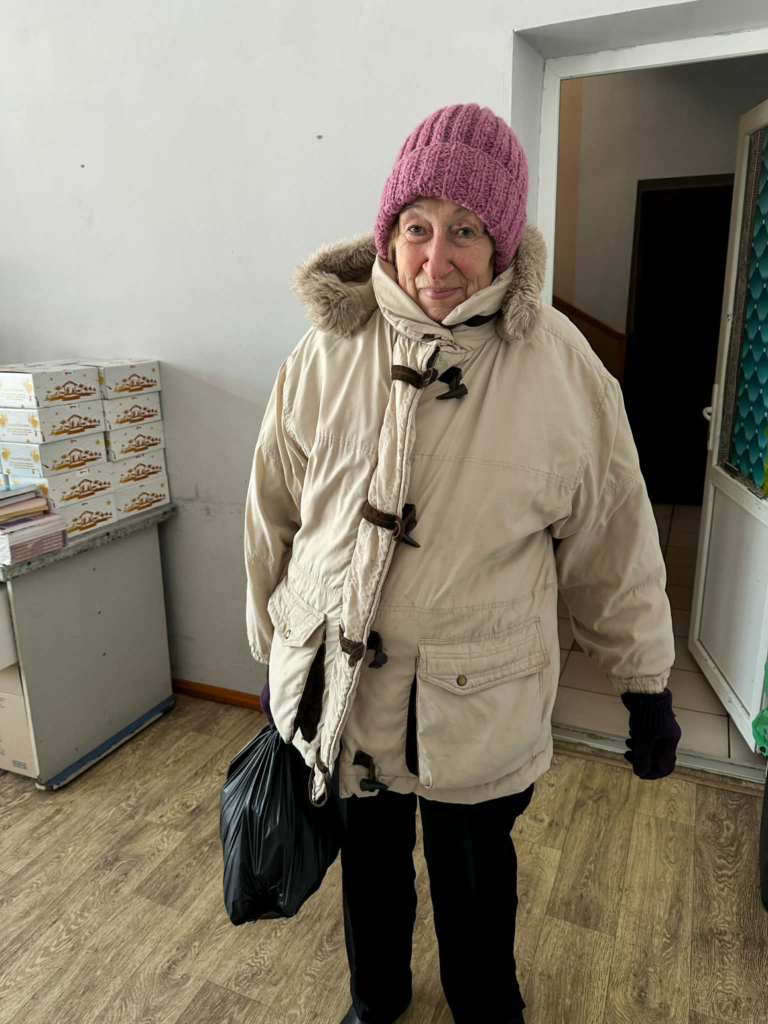 Elderly woman wearing a winter coat and pink knitted hat, holding a bag of aid inside the Centre.
