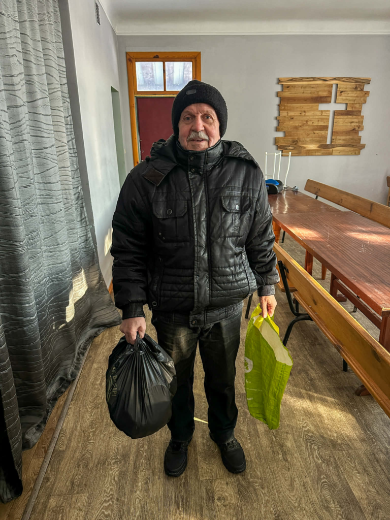 Elderly man in a black winter jacket holding two bags of aid, standing inside the Centre with wooden tables behind him.