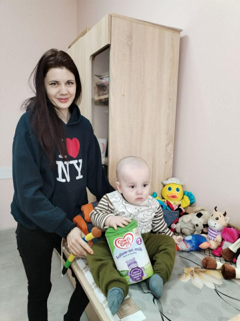 Mother standing beside her baby seated on a table, with baby formula and toys visible, inside the Centre.