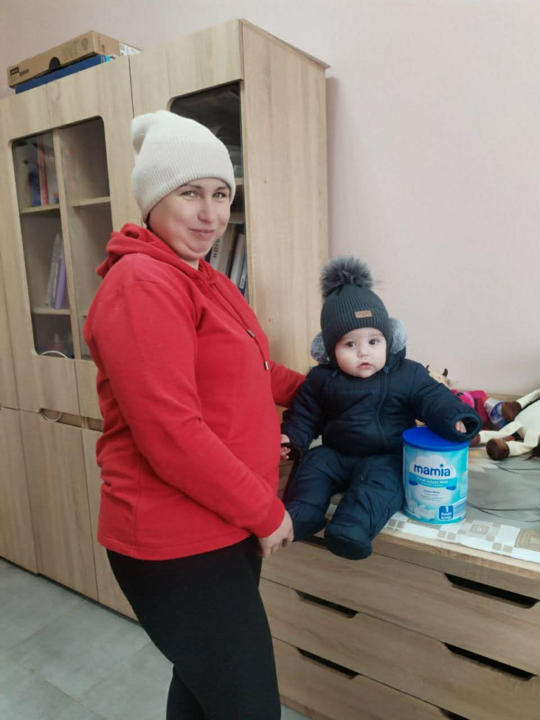 Mother holding her baby dressed in winter clothing, standing next to baby formula on a table in the Centre.