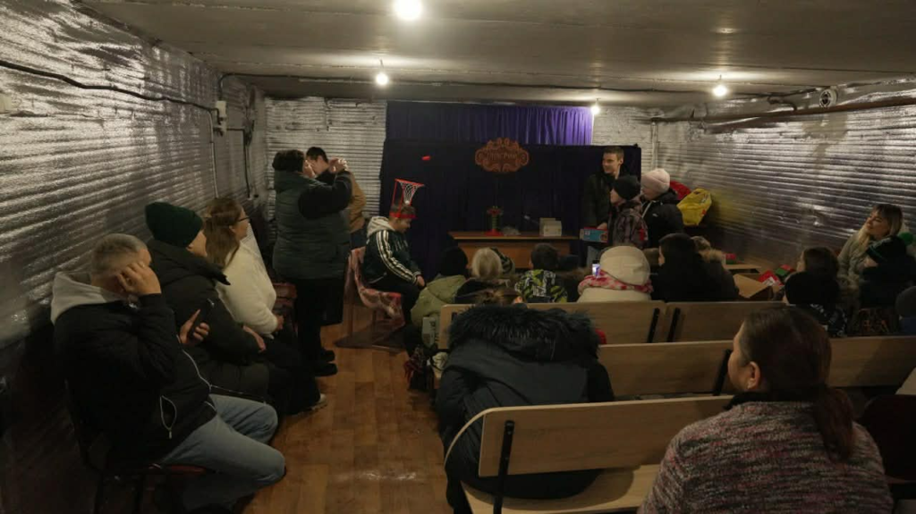 Audience of adults and children seated in the school bomb shelter watching a small puppet show.