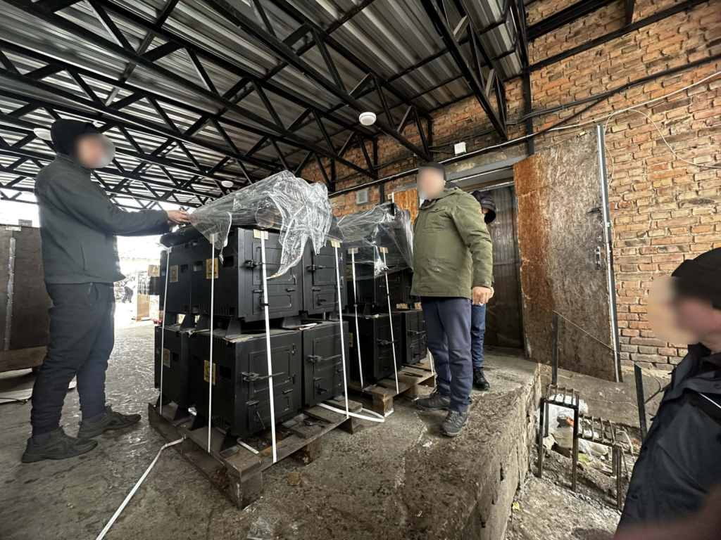 Volunteers stand beside newly manufactured wood-burning stoves secured on pallets inside a warehouse, preparing them for distribution.