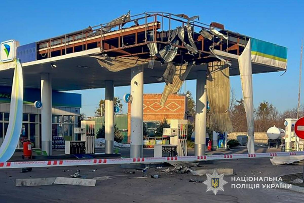 A petrol station canopy severely damaged by a missile strike, with torn metal and debris hanging above the fuel pumps.