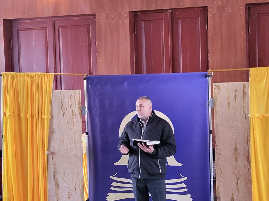 Pastor Andrew speaks while holding a Bible in front of a simple backdrop during a church service in a community hall in Prystaylov.