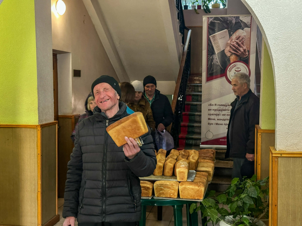 A smiling man holds a loaf of freshly baked bread while others queue behind him to receive food aid indoors.
