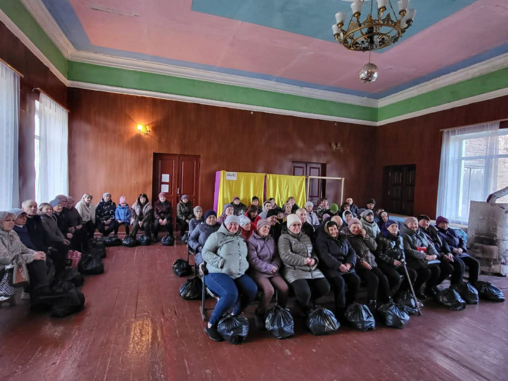 A large group of people sit together in a community hall with bags of aid at their feet during a service in Prystaylov.