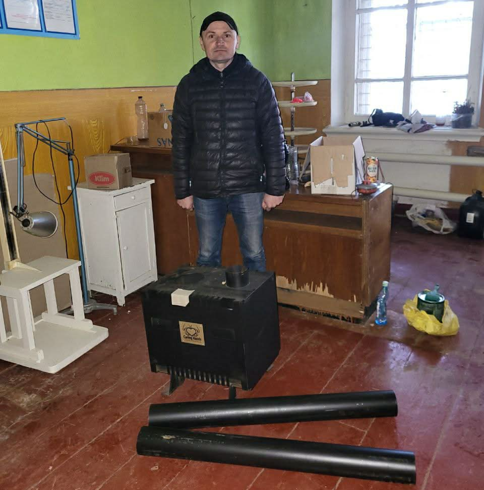 A man stands in a modest room beside a wood-burning stove and flue pipes, prepared for installation to provide heating.