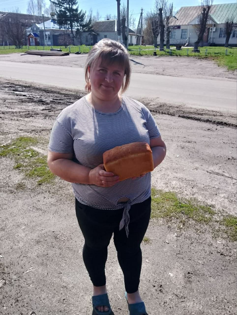 A woman stands outdoors in a village holding a loaf of bread during a local aid distribution.
