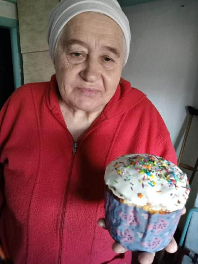 An older woman indoors wearing a red sweater holds a decorated Easter cake topped with white icing and sprinkles.