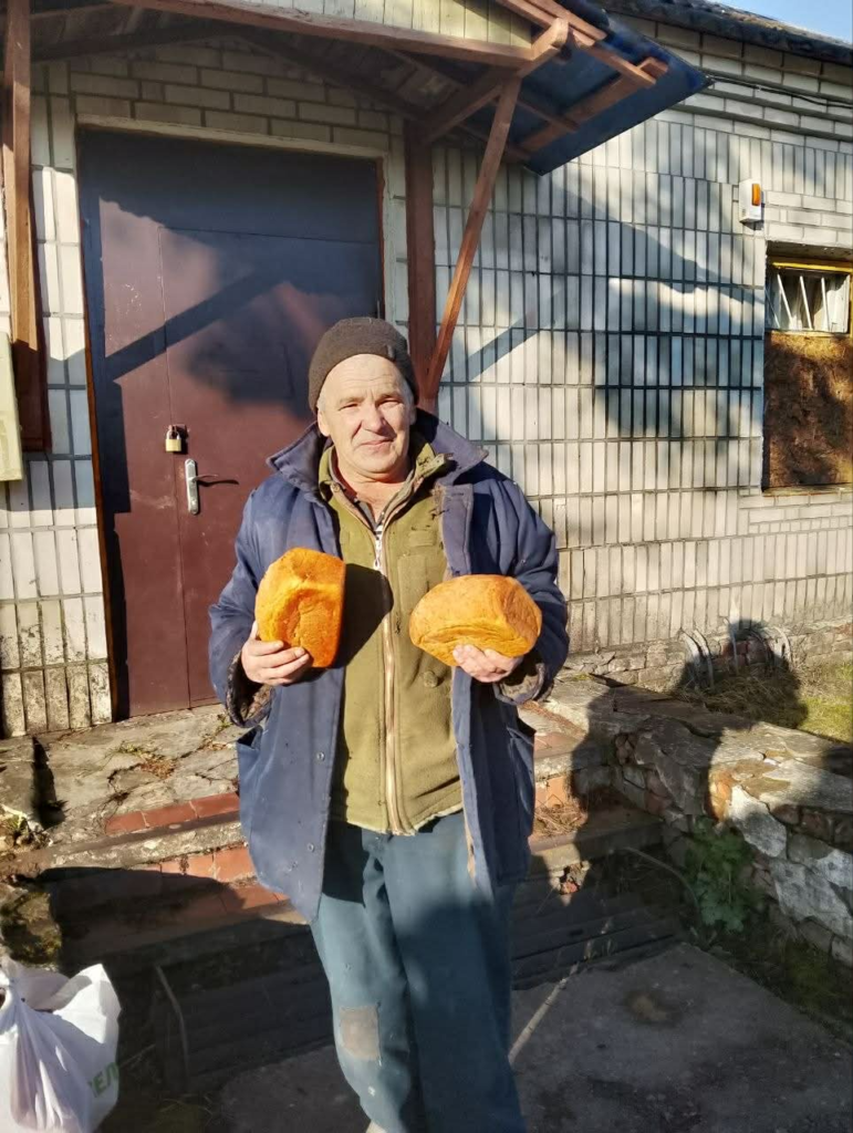 A man stands outside a building holding two loaves of bread received during a village food distribution.