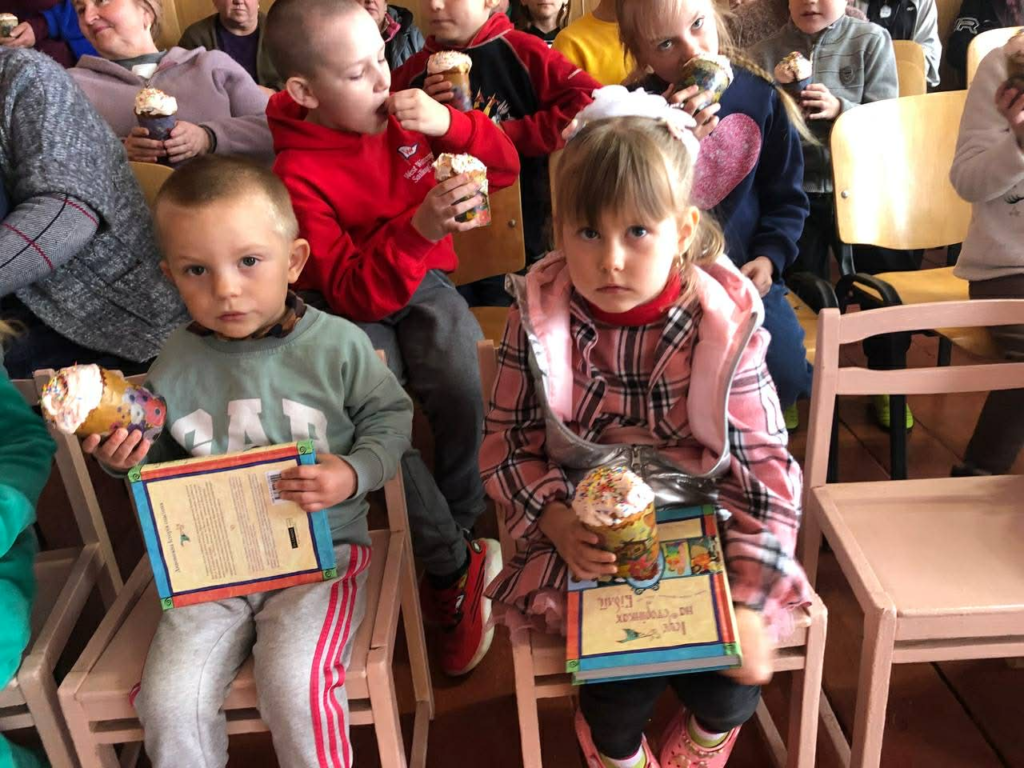 Young children sit on chairs holding decorated cupcakes and Jesus Storybook Bibles, eating and waiting together at an indoor event.