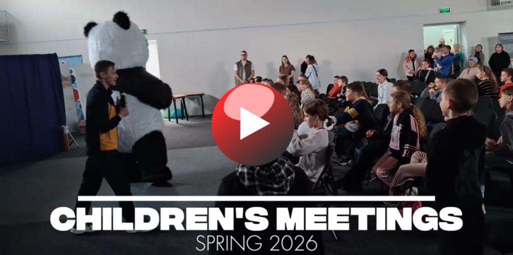 Children seated indoors watching a presenter and a person in a panda costume during a lively Easter 2026 camp meeting..