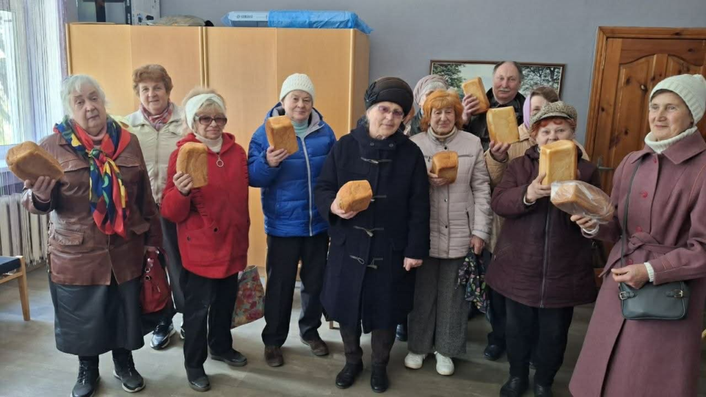 A group of older adults stand indoors holding loaves of bread received during a community aid distribution.
