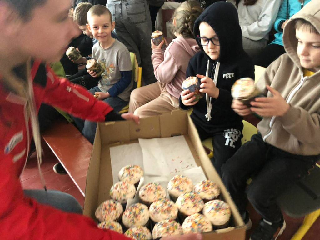 Volunteers distribute decorated cupcakes to children seated indoors during a community outreach event at a school.