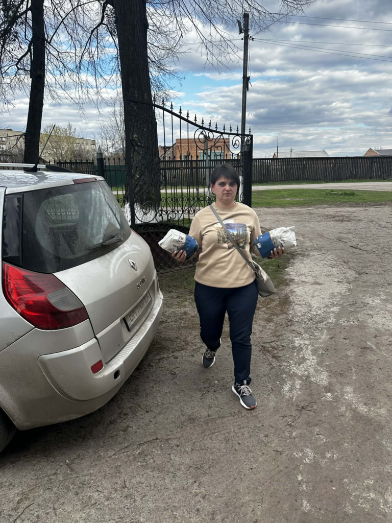 A woman walks beside a parked car carrying two bags of donated flour on a rural road near a gated property.