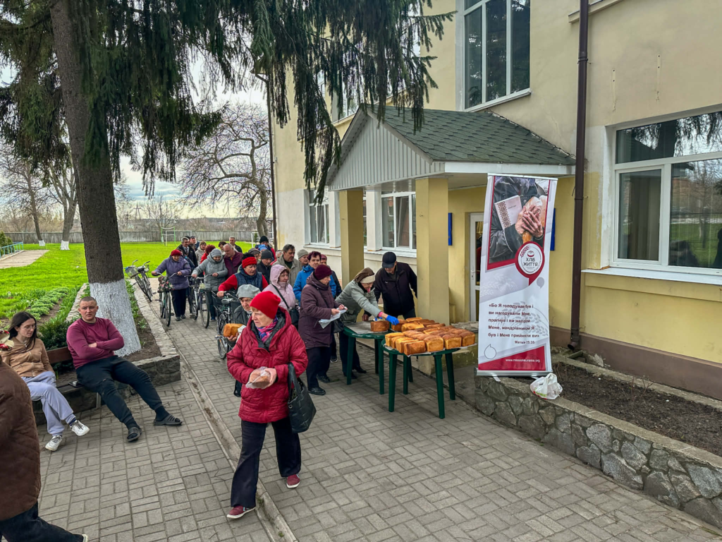 People line up outside the Centre as volunteers hand out fresh loaves of bread from tables.
