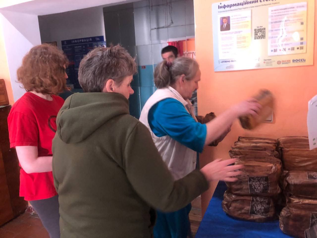 A volunteer sorts and stacks packaged bread during a humanitarian aid distribution inside a community building whilst two ladies wait.