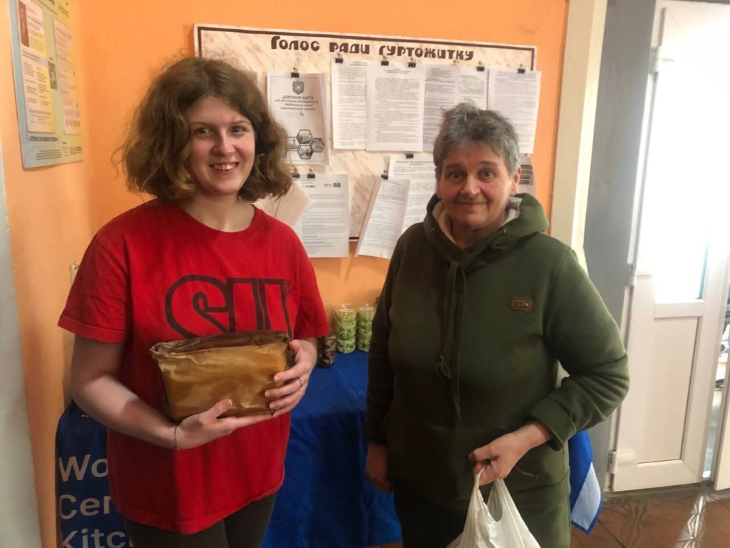 Two women stand indoors during a bread and aid distribution event, at the refugee shelter holding food packages near a community information board.