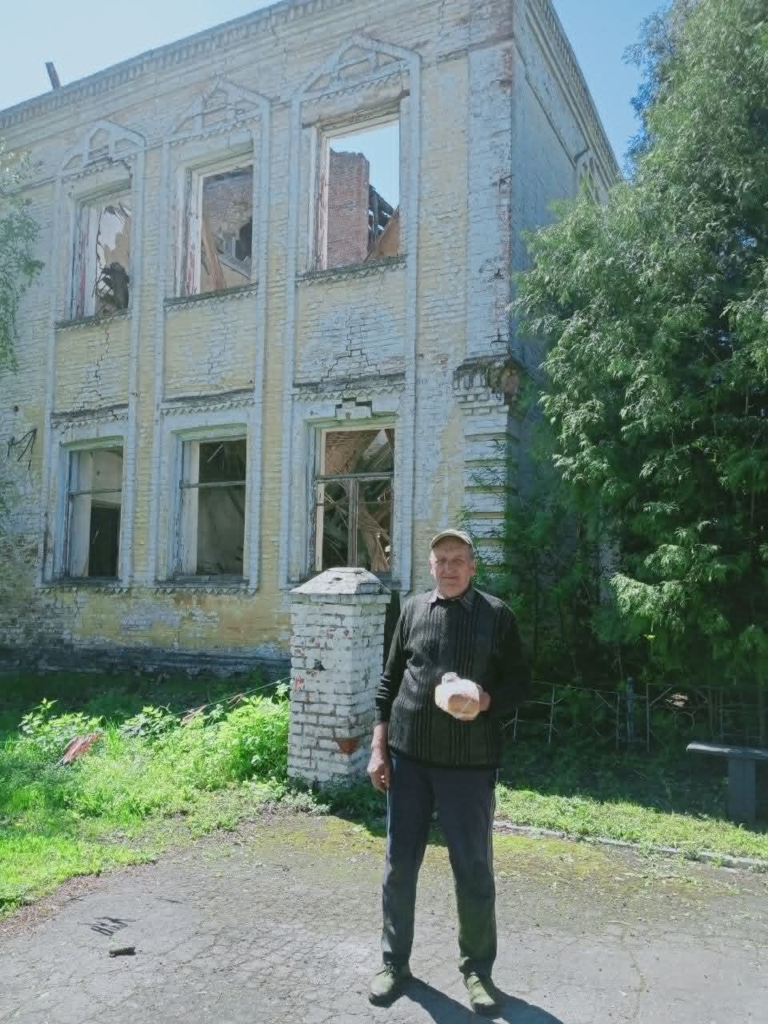 A man stands in front of a heavily damaged building while holding a loaf of bread during a humanitarian aid distribution.
