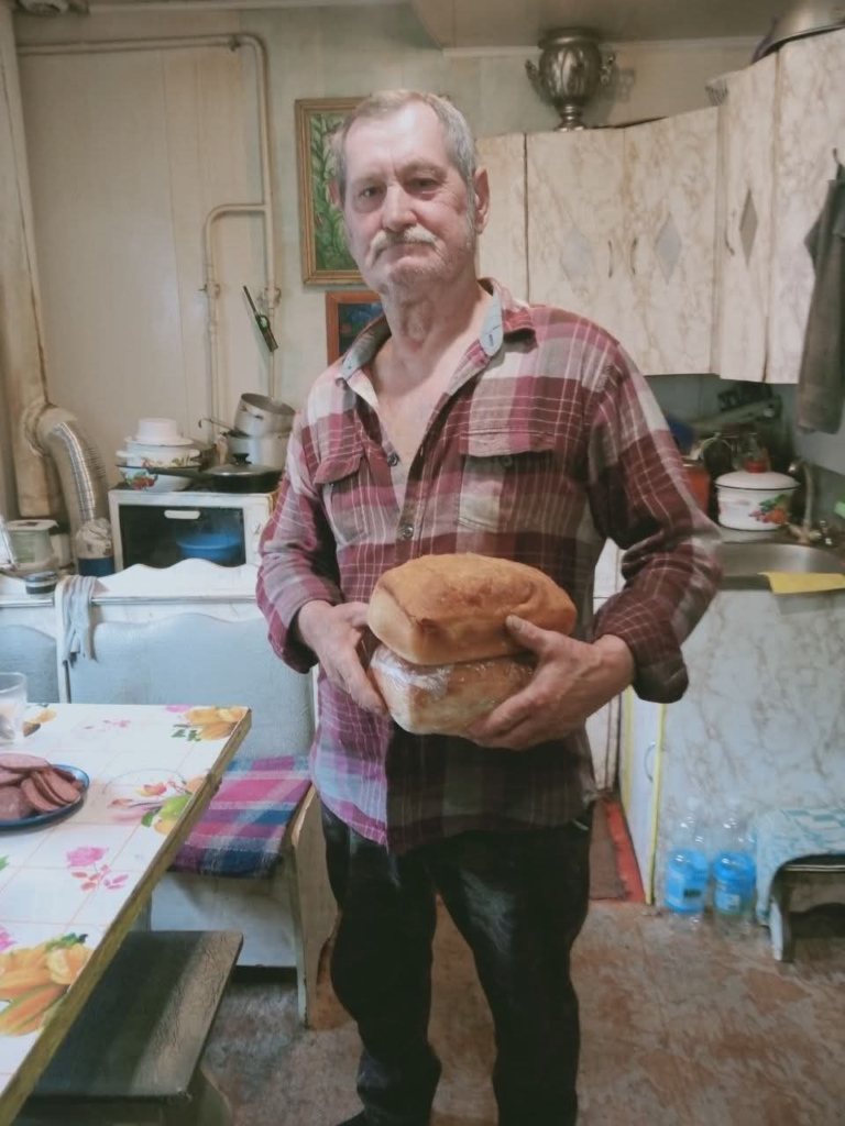 An older man stands in his kitchen holding two loaves of bread received through a food assistance program.