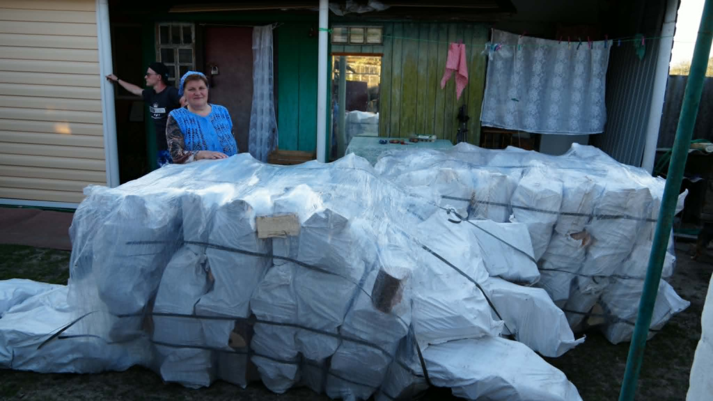A woman and man stand outside their home beside a large delivery of bagged firewood for their wood burner.