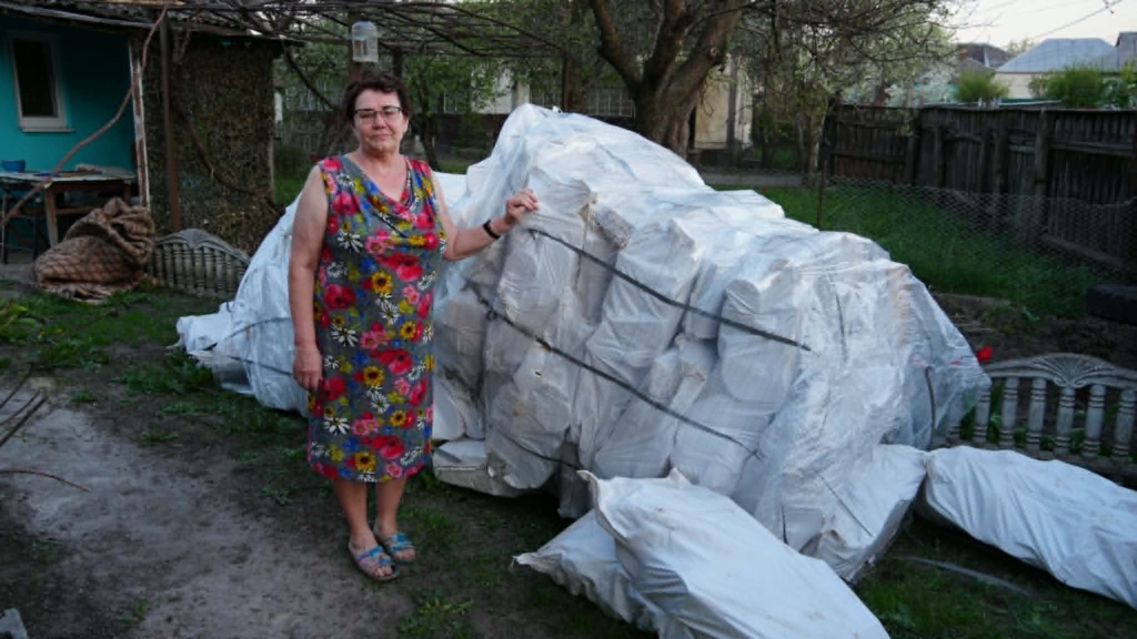 A woman stands in her yard next to a large stack of bagged firewood delivered to her for use in her wood burner.
