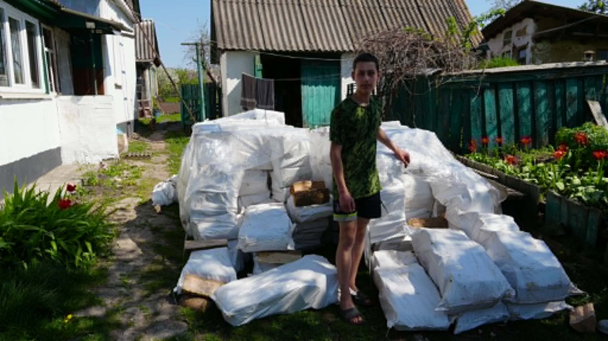 A teenage boy stands beside numerous white bags of firewood stacked outside a rural home.