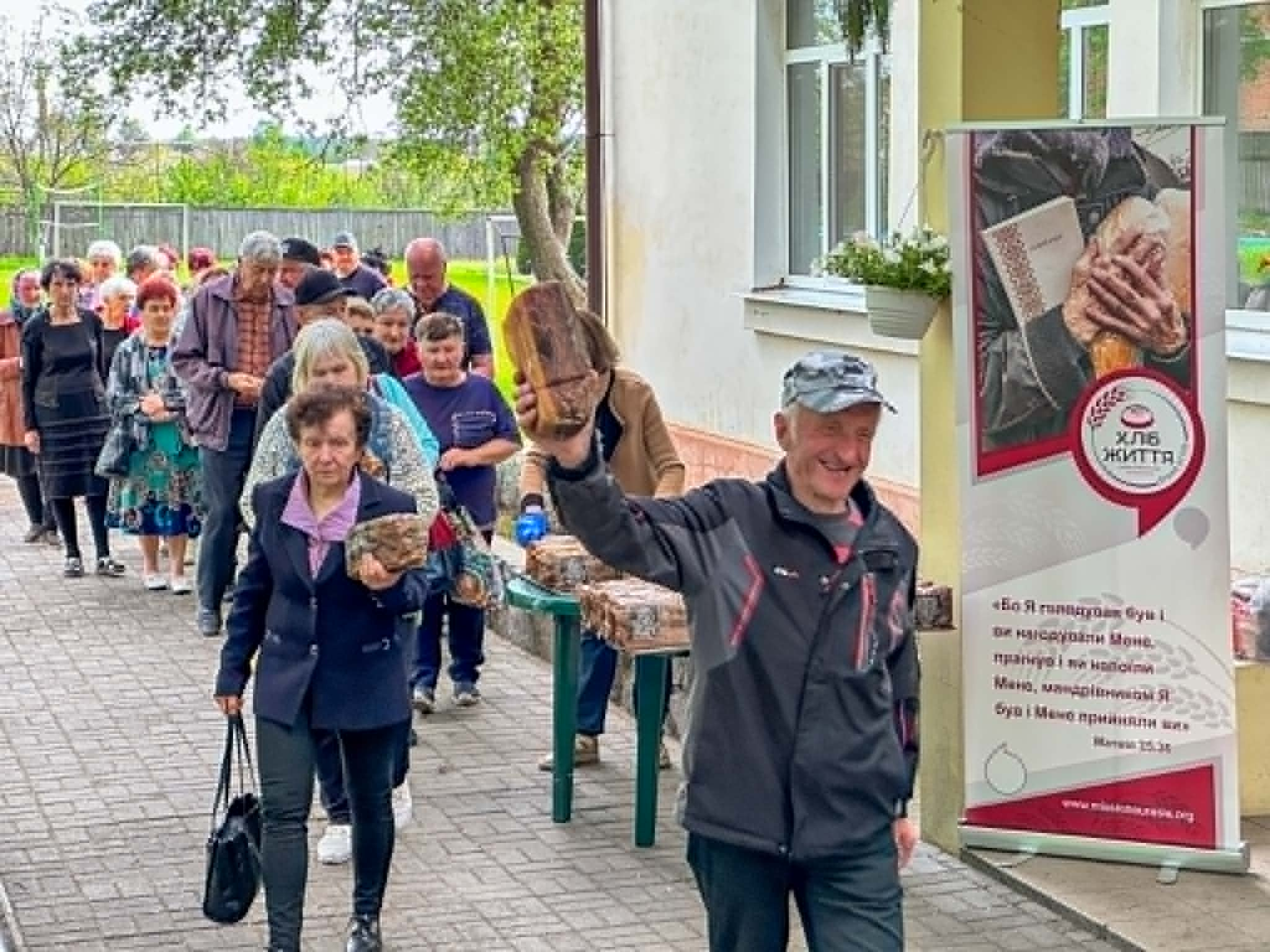 A smiling man lifts a loaf of bread while people line up behind him at a community bread distribution.