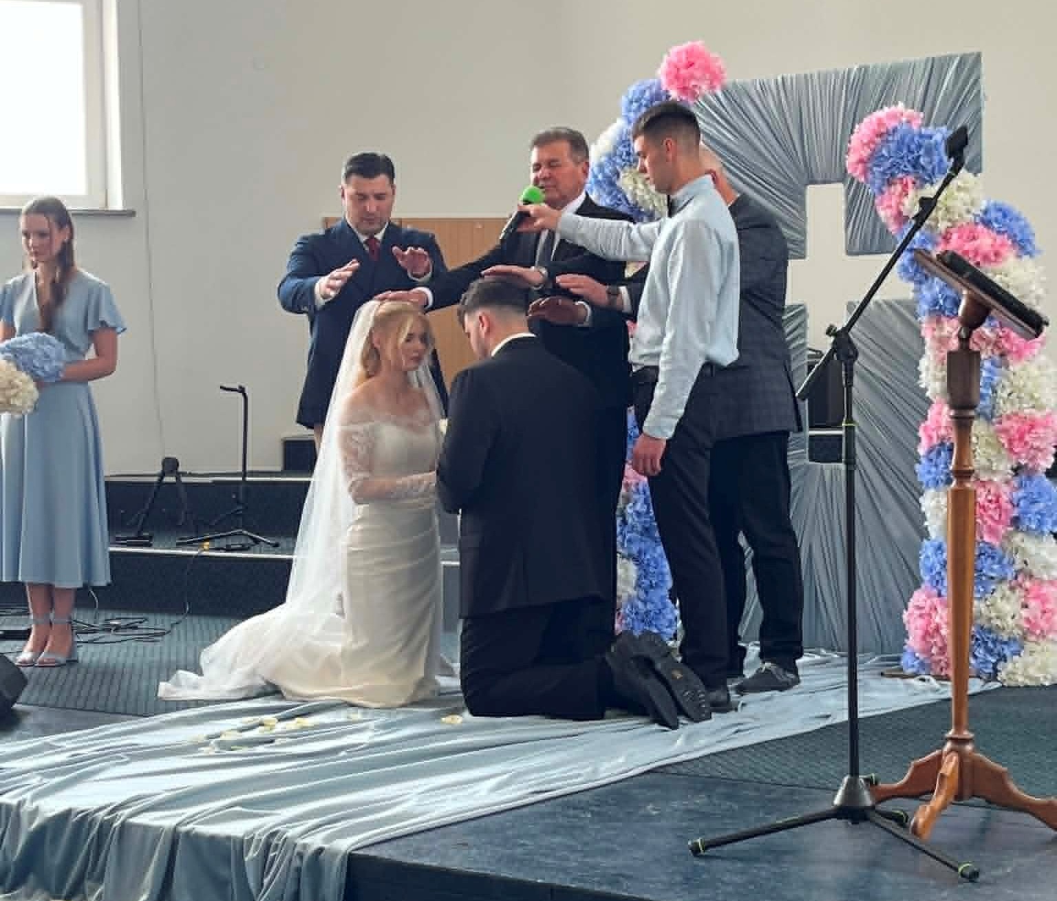 Pastors and family members pray over a bride and groom kneeling during a wedding ceremony inside a church.