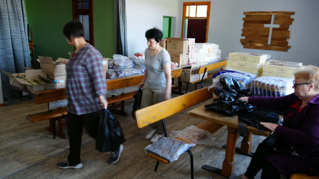 Women organise donated supplies and food packages inside the Centre with benches and stacked boxes in the background.
