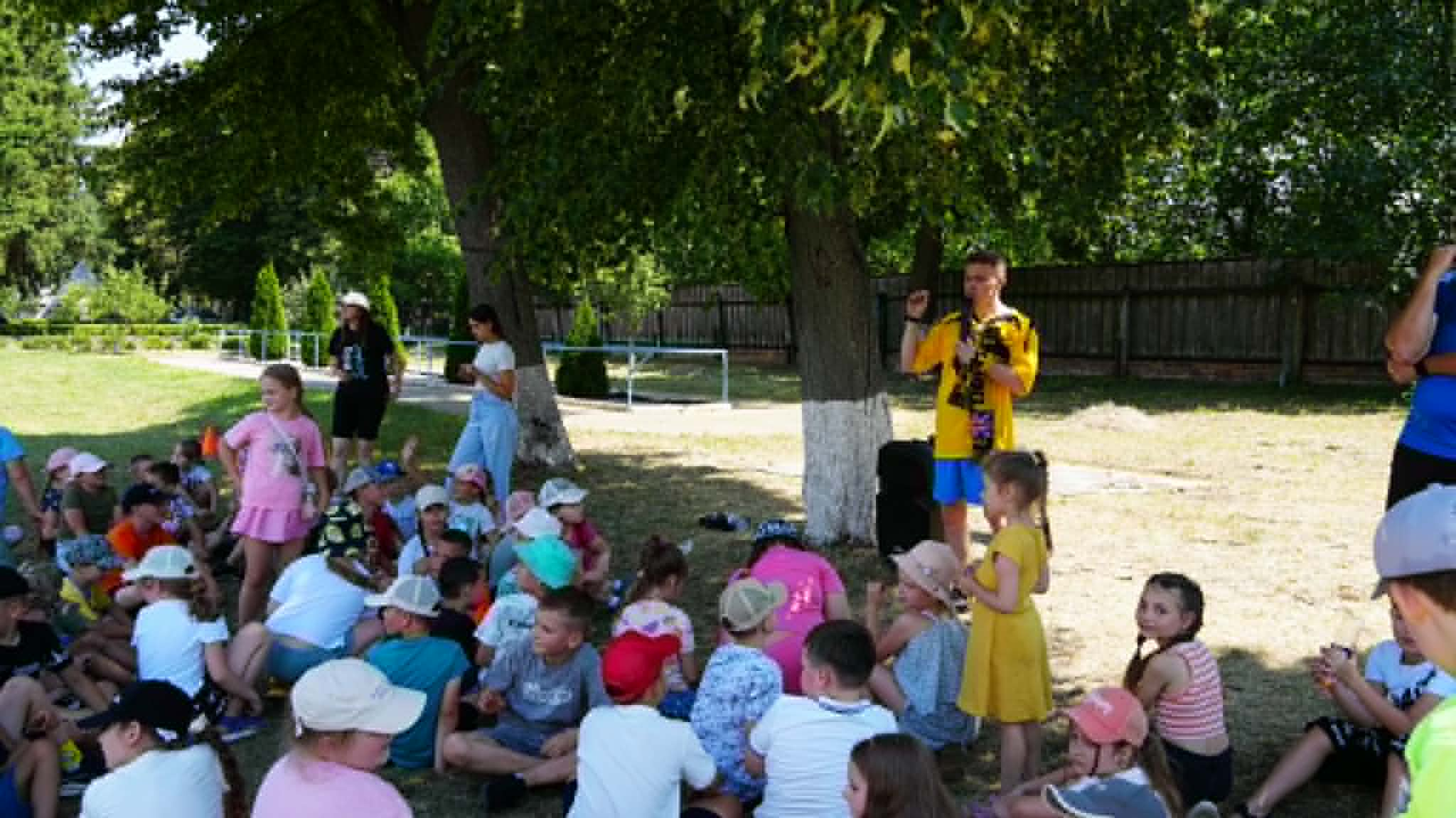 Children gather outdoors under the trees as camp leaders welcome everyone and explain the day's activities at a summer camp.