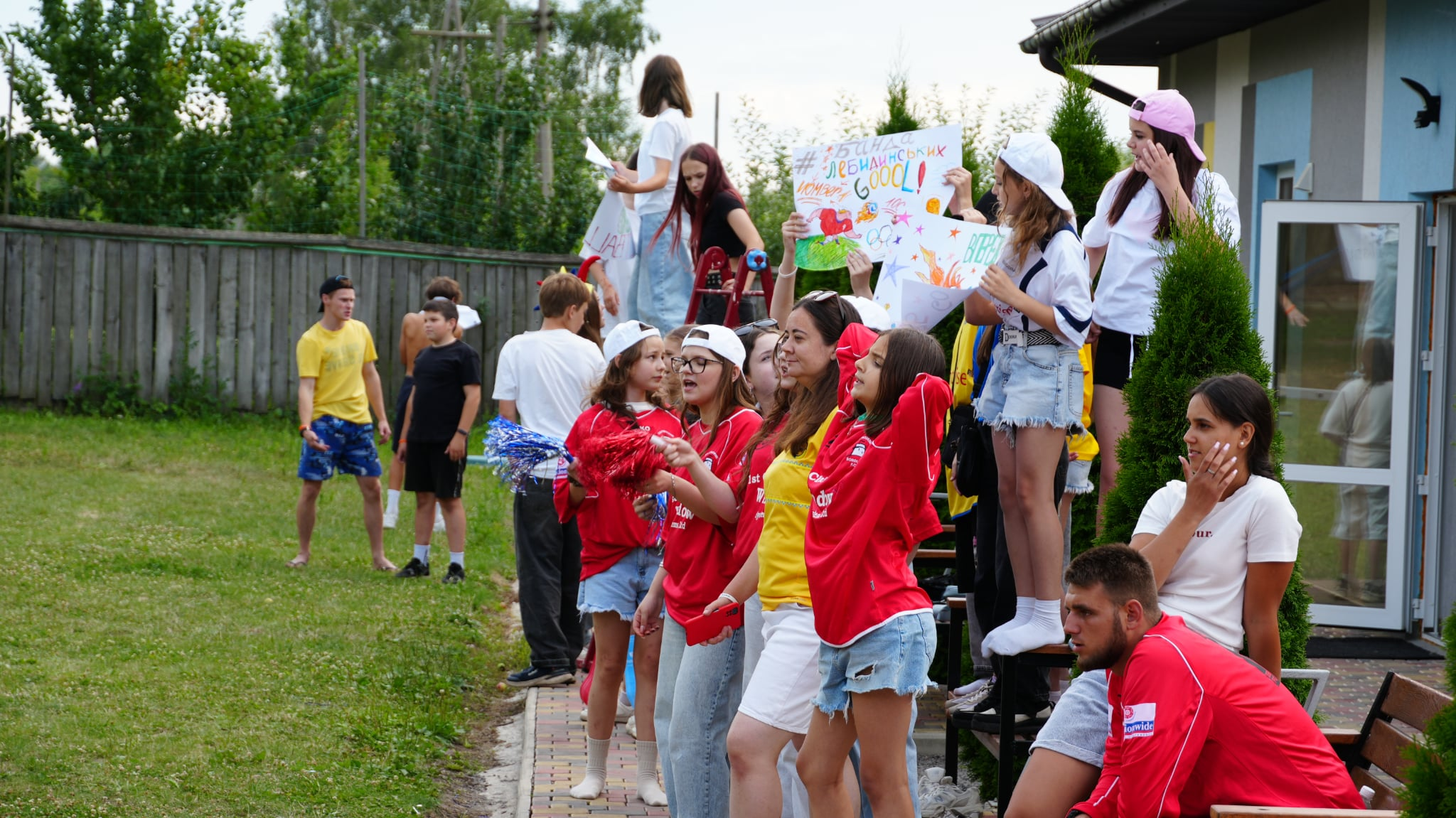 Teenagers cheer on their teammates during an outdoor summer camp challenge, waving pom-poms and handmade banners.
