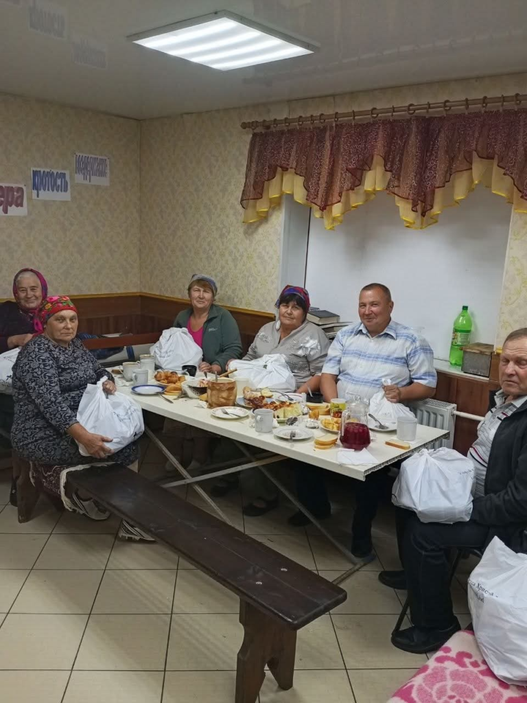 Local people gathered around a table for a Harvest celebration meal, with food aid bags beside them.