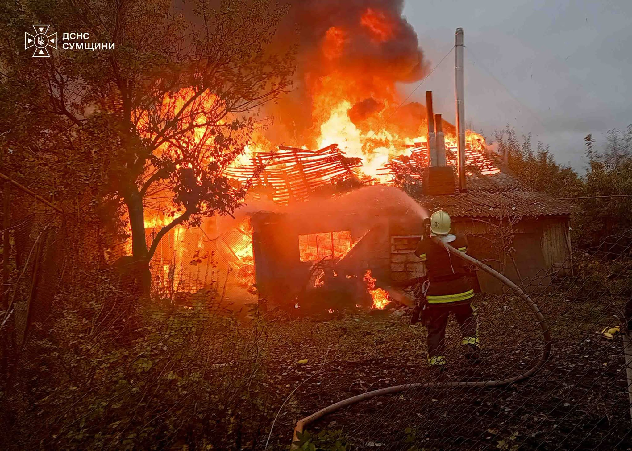 A firefighter tackles a fierce blaze engulfing a house in Lebedyn, with flames and thick black smoke rising above the roof.