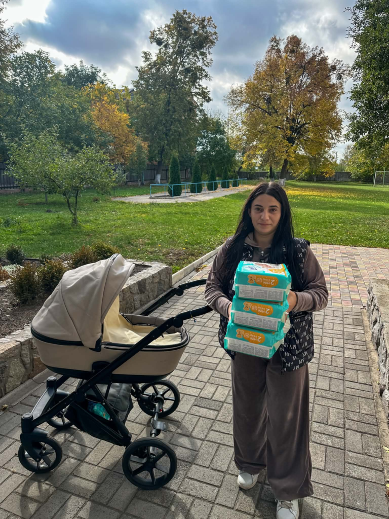 A young Ukrainian refugee stands beside a baby pram, holding three packs of nappies received through the Centre.