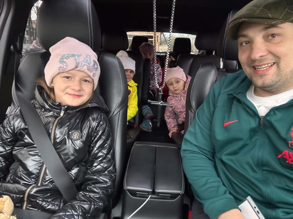Bohdan smiles for a photograph inside a vehicle with several young Ukrainian children dressed in warm winter clothing during a journey to hospital.