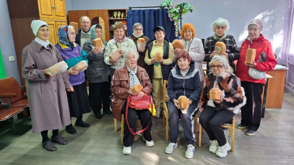 A group of elderly Ukrainian men and women gathered indoors, smiling for a photograph while holding freshly baked loaves of bread.