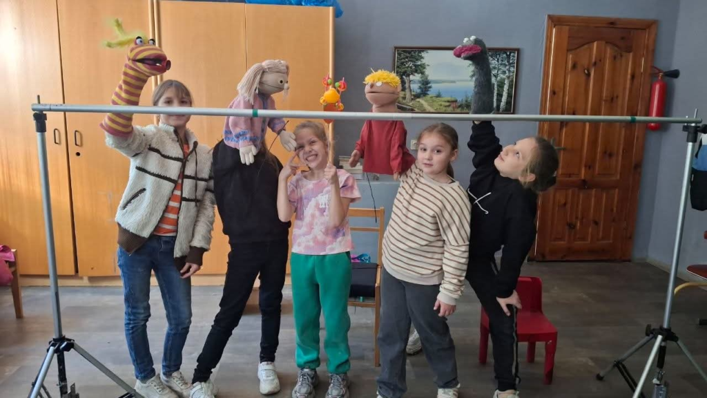 Five Ukrainian children practise a puppet show, holding colourful hand puppets above a makeshift stage during rehearsals.