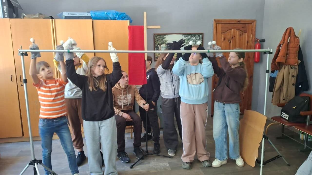 A group of Ukrainian children rehearse a puppet performance, raising animal puppets above a stage while others help behind the scenes.