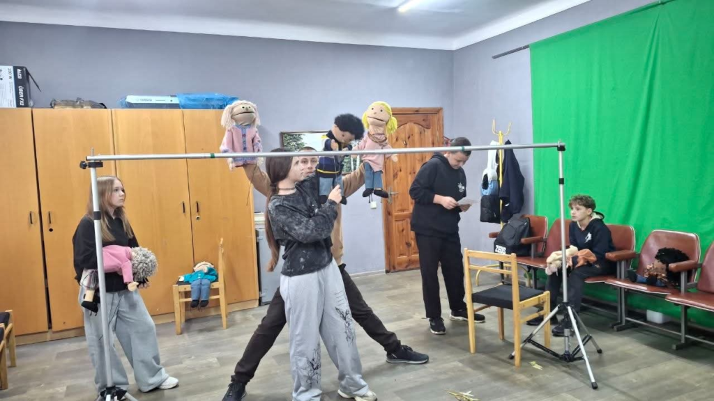 Young Ukrainian puppeteers rehearse with colourful hand puppets in a room equipped with a makeshift stage and green screen.
