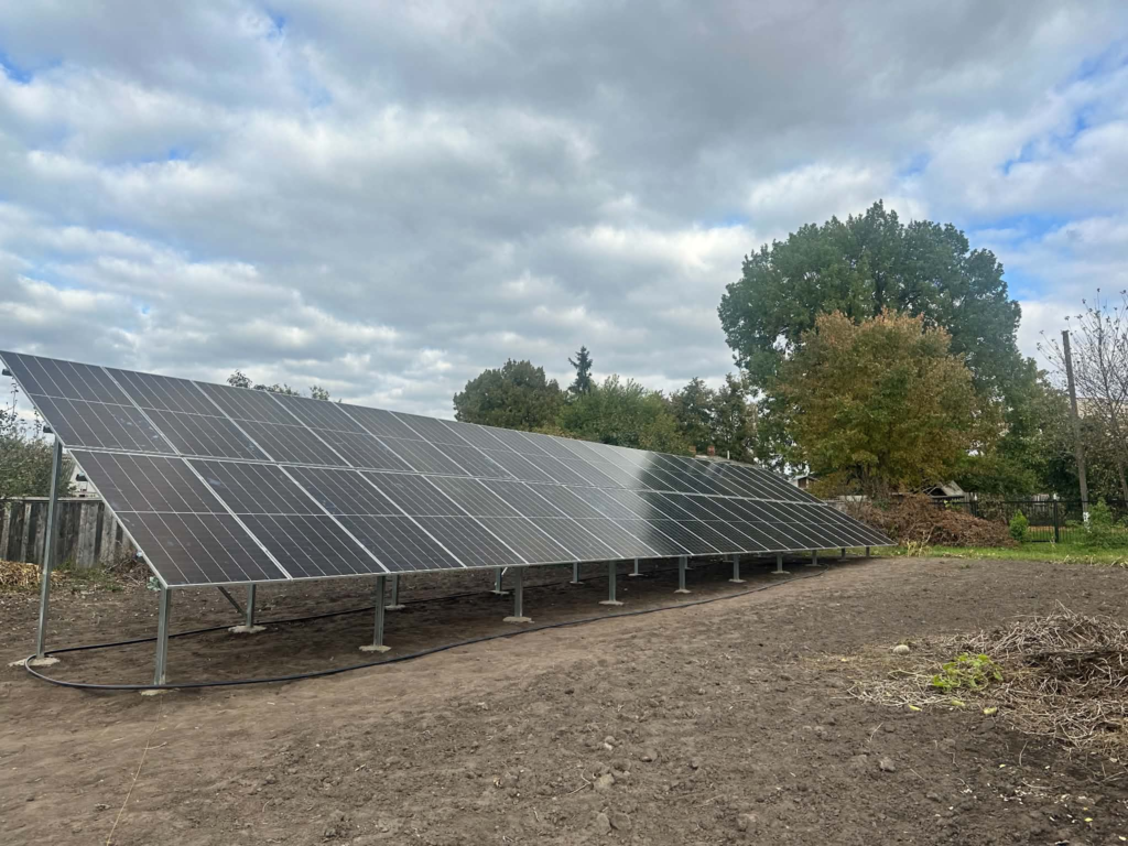 A newly installed array of ground-mounted solar panels in Ukraine, providing renewable electricity, with trees and a cloudy sky in the background.